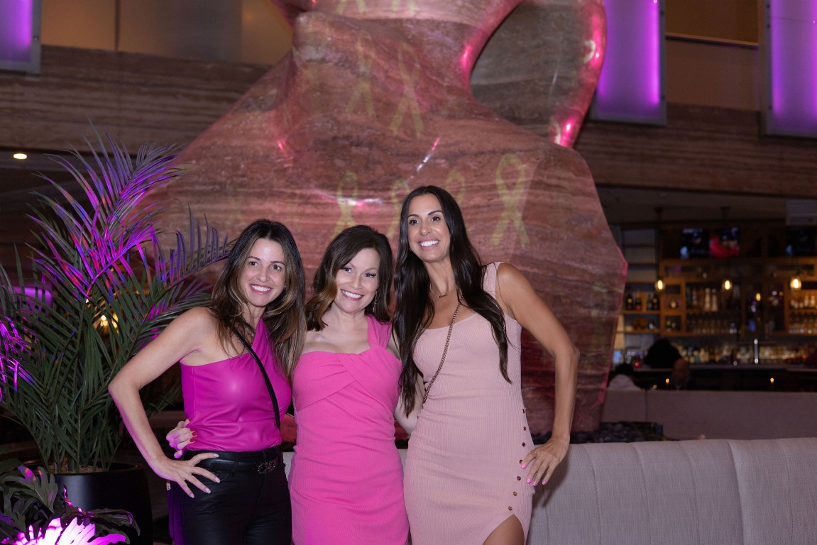 Three women dressed in pink smiling together at a breast cancer awareness event, with pink ribbons projected on the wall behind them